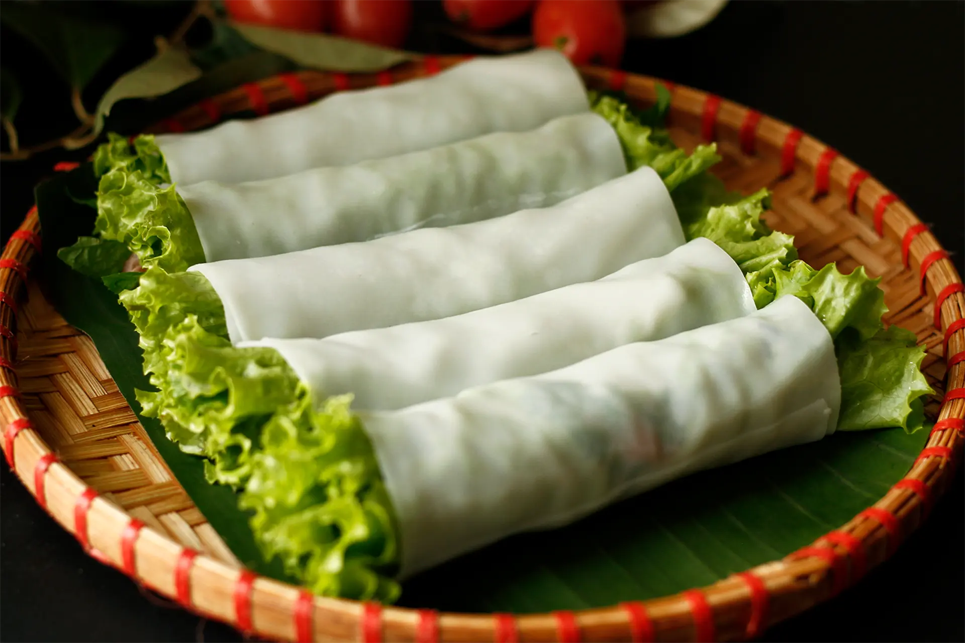 Five phở cuốn rolls — fresh phở sheets wrapped around beef and herbs — served on lettuce and a banana leaf in a red-bound bamboo basket